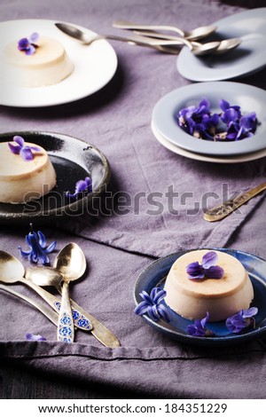 Caramel pannacotta served with violet flowers and vintage spoons over gray textile in retro filter effect.