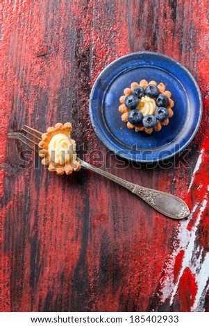 Top view on blueberry mini tart served on blue ceramic plate with vintage fork over black and red wooden background.