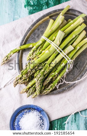 Bunch of young green asparagus on vintage tray served with ceramic plate of sea salt over green wooden table. Top view.