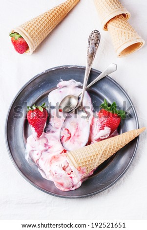 Wafer cones with strawberry ice cream with syrup, spoons and fresh strawberries served on vintage plate over white textile. Top view.