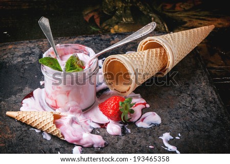 Strawberry ice cream with mint and fresh strawberries served in glass jar with empty wafer cones over dark table.