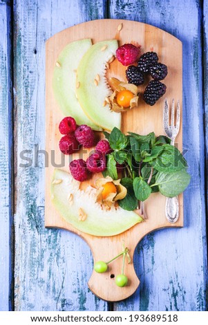 Mix of raspberries, blackberries, mint, physalis and melon on wooden cutting board over purple wooden table. Top view.