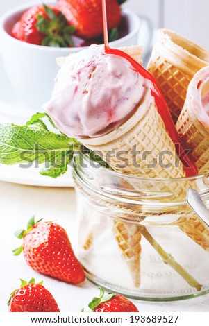 Wafer cones with strawberry ice cream with pouring syrup and fresh strawberries served in glass jar over white textile.