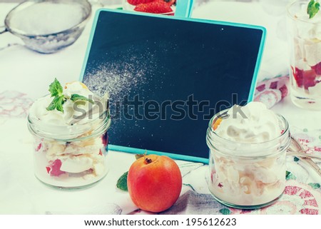 Dessert Eton mess with merengue, berries and whipped cream, served in glass jars on table with empty chalkboard in instagram effect