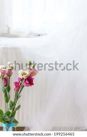 Fragment of interior with bouquet of fresh pink eustoma flowers  in blue vase with window and white curtains as background