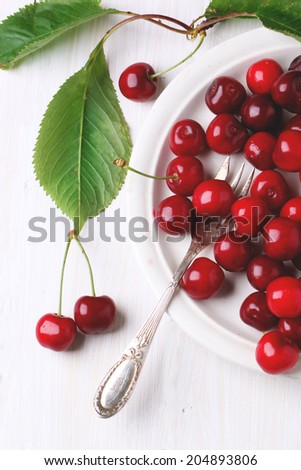 Ceramic plate of fresh cherries with leaves, served with dessert fork over white wooden table. Top view.