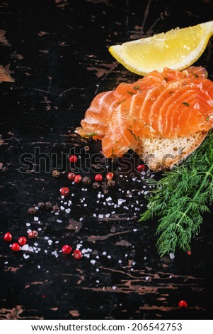 Bread with sliced salted salmon, served on black wooden table with lemon, sea salt and peppers.