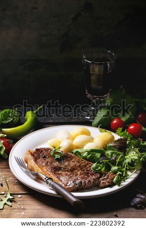 Grilled steak with potatoes and green salad on white ceramic plate and vintage glass with red wine over old wooden table. See series