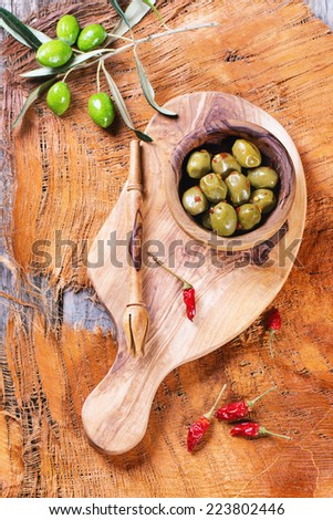 Green olives in olive wood bowl with chili pepper and olive\'s branch served on cutting board over wooden table. Top view.