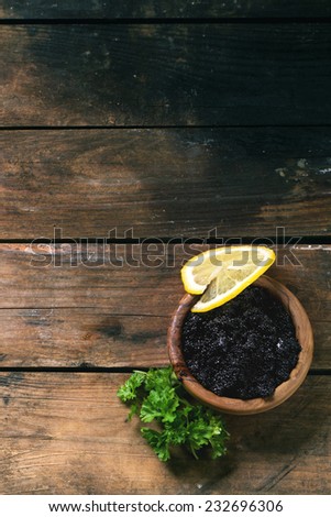 Wooden bowl of black caviar with lemon slice and parsley over old wooden table. Top view.