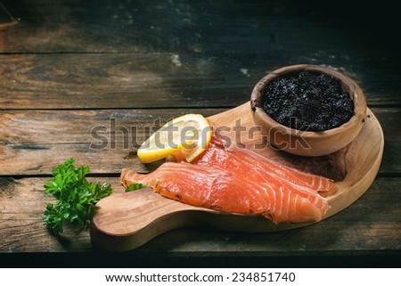 Bowl of black caviar and pieces of salted salmon on olive wood board with vintage knife over old wooden table