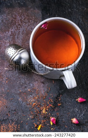 Vintage aluminum mug of hot tea with tea strainer, dry tea and rose buds over dark background. Top view