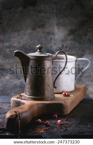 Vintage teapot and aluminum mug of tea, served on wooden cutting board with dry rose buds over dark background