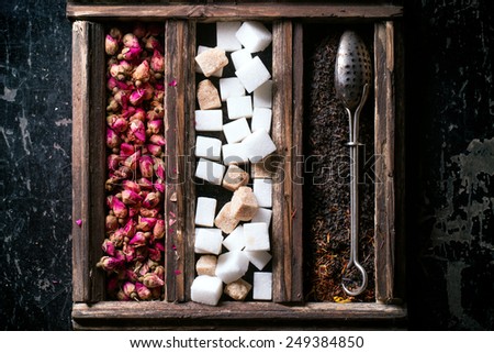 Set of dry black tea, tea roses and sugar cubes in wooden box with vintage tea strainer over black backgrounda