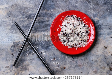 Uncooked black and white rice in red plate with black wooden chopsticks over tin surface. Top view.
