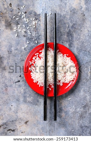 Uncooked rice in red plate with black wooden chopsticks over tin surface. Top view.
