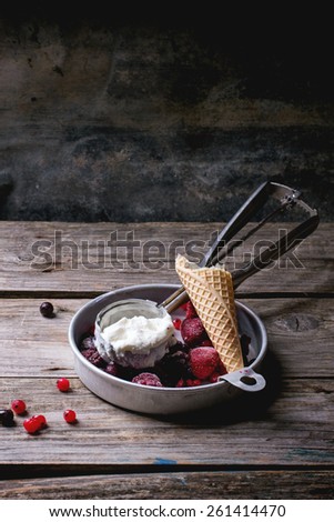 Aluminium plate with homemade vanilla ice cream, mix of frozen berries and bitten waffer cone, served with metal spoon over wooden table
