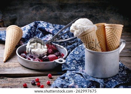Homemade vanilla ice cream in wafer cones and empty waffer cones, served in metal bowl with frozen berries and spoon over wooden table with blue textile
