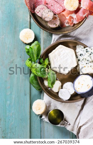 Metal plates with cheese and sausage variations with fresh basil and olive oil over turquoise wooden table. Overhead view.