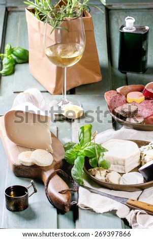Plates with cheese and sausage variations with fresh basil and glass of white wine over turquoise wooden table. Overhead view.