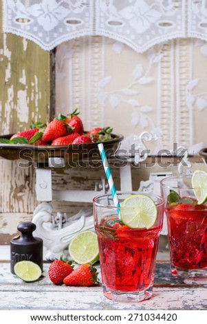 Two glasses with retro cocktail tubes and glass jug of homemade strawberry lemonade, served with fresh strawberries, mint, lime and ice cubes over old white wooden table. See series