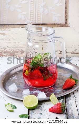Glass jug of homemade strawberry lemonade, served with fresh strawberries, mint, lime and ice cubes over old white wooden table. See series