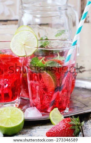 Two glasses with retro cocktail tubes and glass jug of homemade strawberry lemonade, served with fresh strawberries, mint, lime and ice cubes over old white wooden table. See series