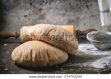 Two homemade wholegrain pita bread on wooden cutting board, served with vintage rolling-pin and flour over dark table