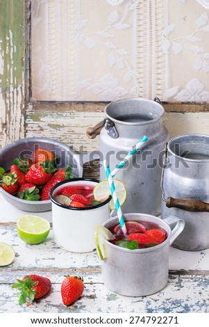 Vintage metal cups with retro cocktail tubes and water-cans of homemade strawberry lemonade, served with fresh strawberries, mint and lime over old white wooden table. See series