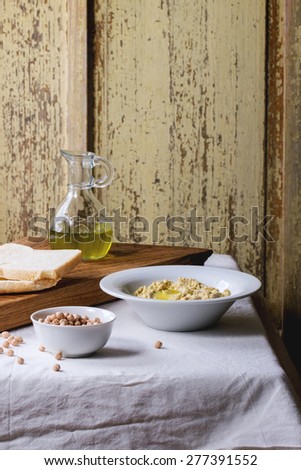 Plate of homemade hummus with bread, olive oil and raw chick-pea, served on wooden cutting board over rustic table with gray tablecloth