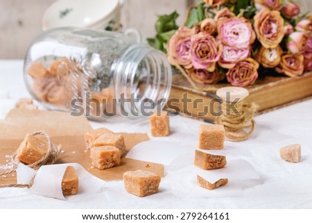 Fudge candy and caramel on baking paper and in glass jar, served over white tablecloth with bouquet of dry pink roses and thread for gift making. See series