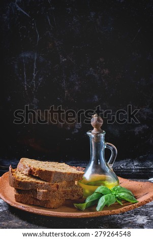 Clay plate woth sliced homemade rye bread, bottle of olive oil and fresh basil over black powdered with flour table.