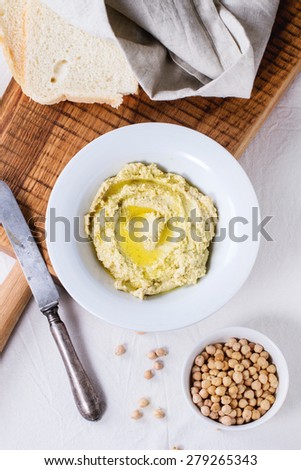 Plate of homemade hummus with bread, olive oil and raw chick-pea, served on wooden cutting board over rustic table with gray tablecloth. Top view