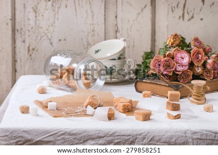 Fudge candy and caramel on baking paper and in glass jar, served over white tablecloth with bouquet of dry pink roses and thread for gift making. See series