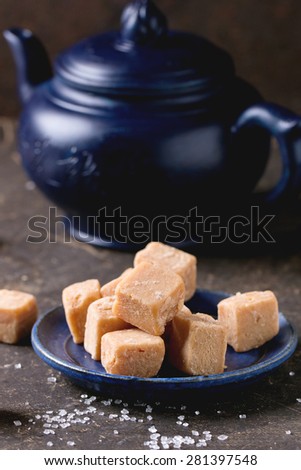 Blue ceramic plate with fudge candy ceramic teapot, served with sugar cubes over dark background.