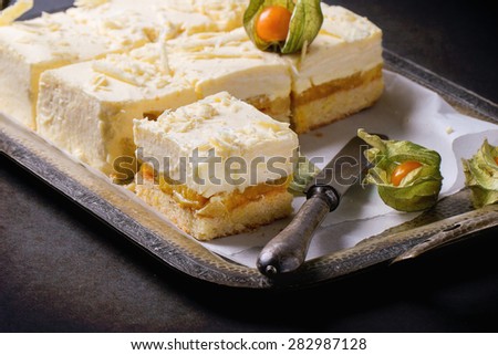 Homemade sliced cake with creamy mousse and tropical fruits mango and physalis served in vintage metal tray over black background