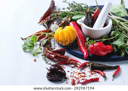 Assortment of fresh, dryed and flakes hot chili peppers and fresh herbs with white ceramic mortar on dark blue cutting board over light blue wooden background. Selective focus.