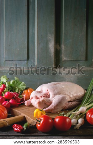 Raw young duck on wooden cutting board with fresh ripe vegetables over wooden table. Dark rustic atmosphere.