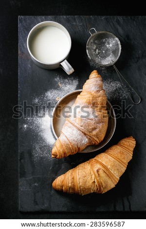 Two fresh baked croissants with sugar powder served with aluminum cup of milk and vintage sieve over black slate background