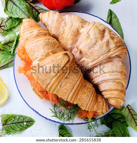 Croissant sandwich with salted salmon on white plate, served with fresh salad leaves, lemon, sea salt and vegetables over light blue wooden background. Top view. Square image