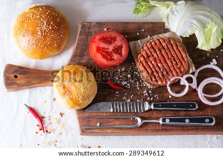 Ingredients for making homemade burger on wooden cutting board, served with meat fork and knife over White tablecloth. Dark rustic style. Top view