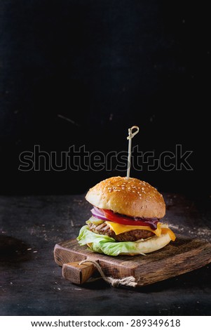 Fresh homemade burger on little wooden cutting board over dark background.