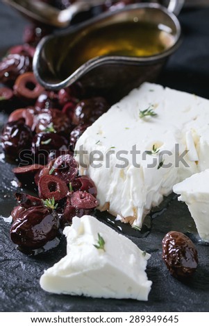 Whole and sliced black olives and block of feta cheese with olive oil on black slate over dark background. Selective focus