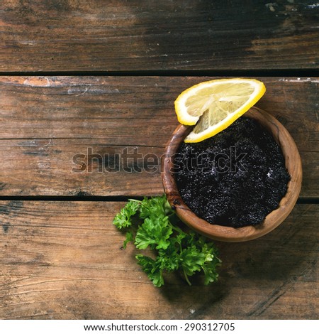 Wooden bowl of black caviar with lemon slice and parsley over old wooden table. Top view. Square image