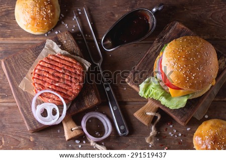 Fresh homemade burger on little cutting board and raw cutlet and sliced onion, served with ketchup sauce and meat fork over wooden table. Dark rustic style. Top view