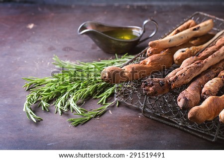 Fresh baked homemade grissini bread sticks in vintage metal grid box with olive oil and herbs rosemary and thym over dark surface.