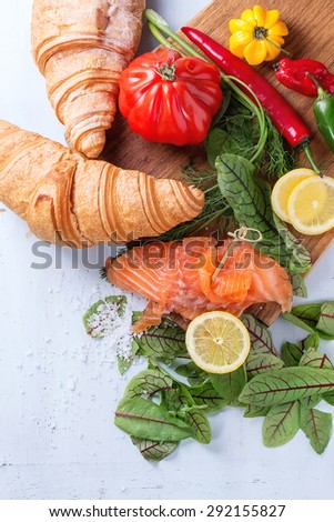 Slaced salted salmon with fresh salad leaves, lemon, sea salt and vegetables, served on wooden chopping board with croissant over light blue wooden background. Top view.