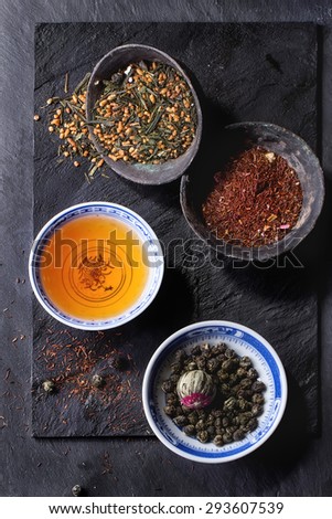 Assortment of dry tea and cup of hot tea. Green tea, black tea, green tea with rice, rooibos, dry rose buds in porcelan and old metal chinese bowls. Black textured slate background. Top view.