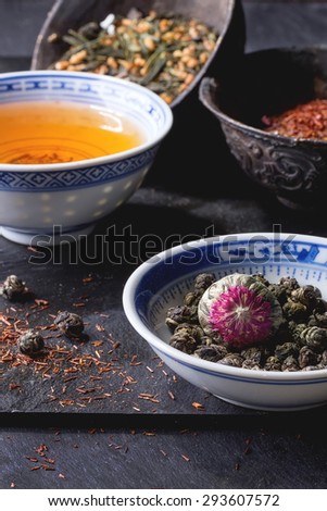 Assortment of dry tea and cup of hot tea. Green tea, black tea, green tea with rice, rooibos, dry rose buds in porcelan and old metal chinese bowls. Black textured slate background
