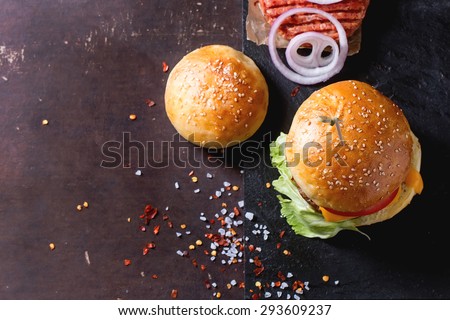 Fresh homemade burger on black slate and raw cutlet and sliced onion, served with sea salt and pepper over dark background. Top view. With copy-space on left.